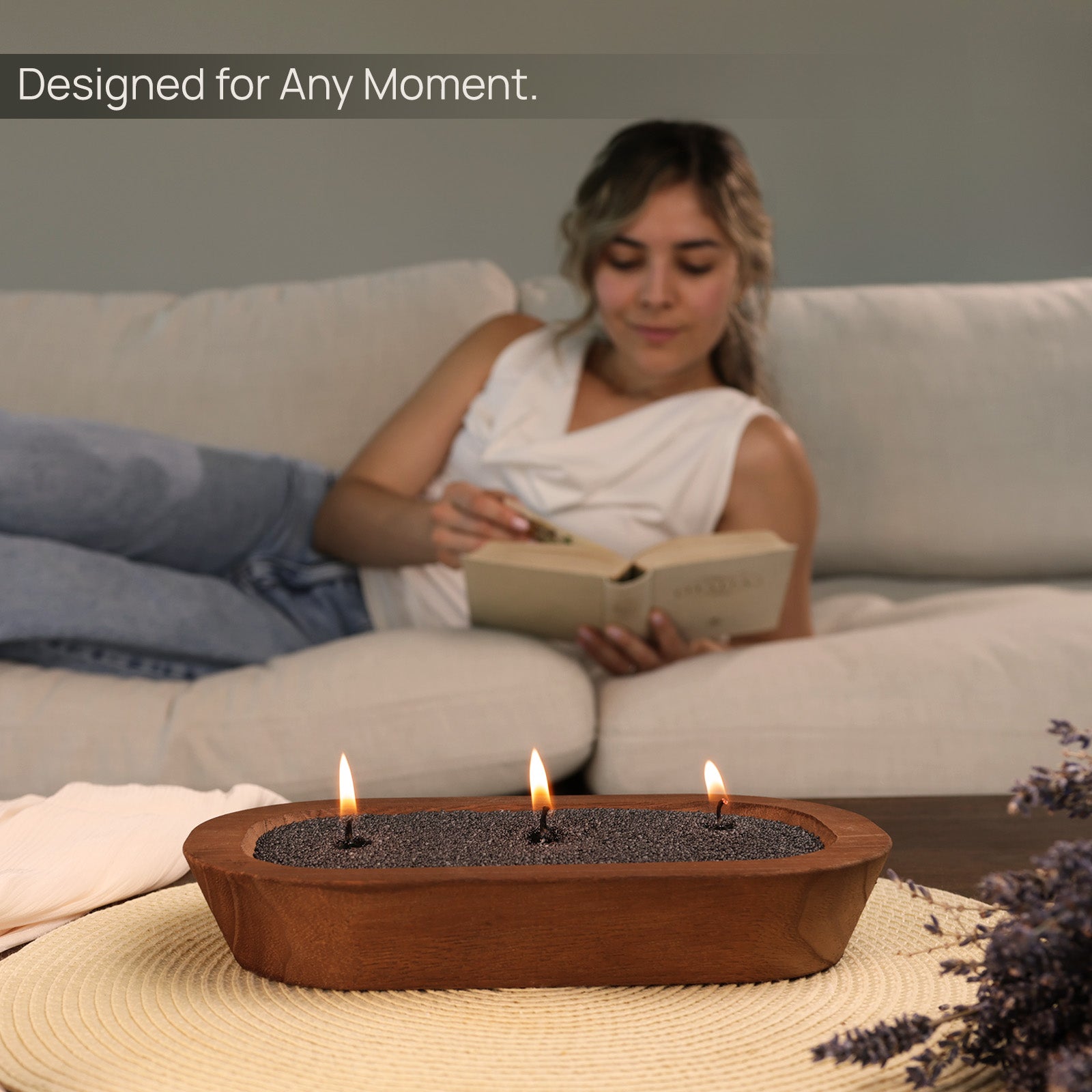 Woman relaxing on a sofa with a book, while a Tula black sand candle burns in a wooden bowl on a woven mat. Cozy, calming ambiance designed for self-care, mindfulness, evening routines, and intentional living. Text overlay reads “Designed for Any Moment.